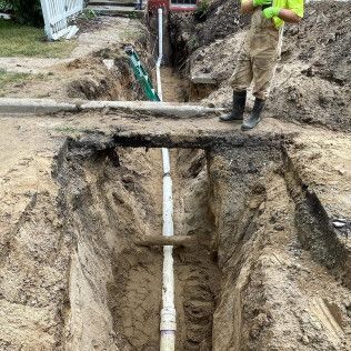 A trench dug with a white pipe running through it, a worker standing nearby.