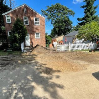 Brick house with dirt pile in front. White picket fence, small house, and trees in background.