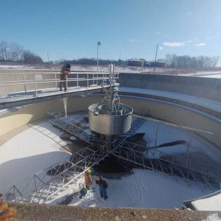 Workers in a wastewater treatment tank. One on walkway, two on a metal structure. Snow on the water, clear sky.