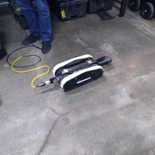 A small, white and black robotic device with treads on a concrete floor with a person in jeans standing nearby.