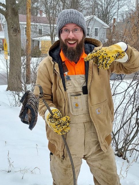 Man in Carhartt jacket and overalls, covered in mud and snow, holds a muddy stick and points to his gloves.