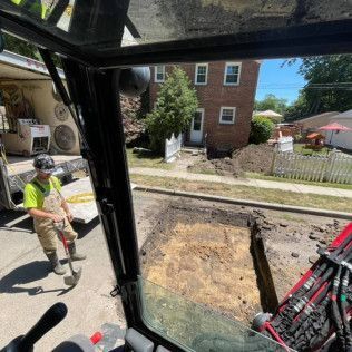 Worker stands near excavation in street. View from inside construction equipment. Buildings in background.