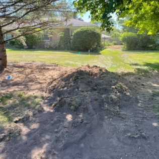 Mound of dirt in a yard, with a house and trees in the background on a sunny day.