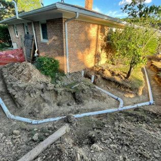 Construction site with white drainage pipes laid in trenches around a brick house. Soil piles visible.