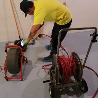 Plumber in a yellow shirt inspects a drain with a camera, using tools in a basement.