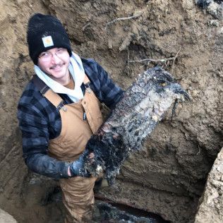 Man in overalls and a beanie holds a large, dark, weathered piece of material while standing in a trench.