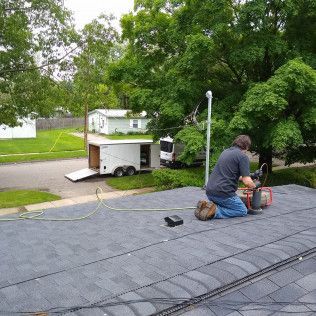Man on a roof repairs shingles; trailer and truck parked in the street nearby.