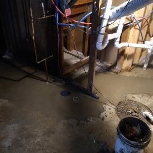Basement with standing water, pipes, exposed wall studs, and a bucket.