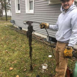Man holding long, dark, tangled debris extracted from a drain; outdoors, beside a house.