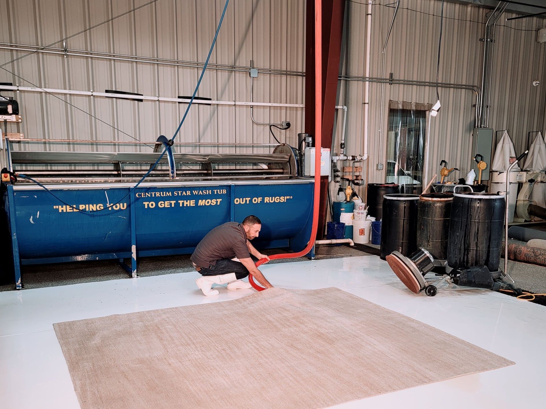 Worker in a warehouse spreads coating on the floor beside blue industrial tanks.
