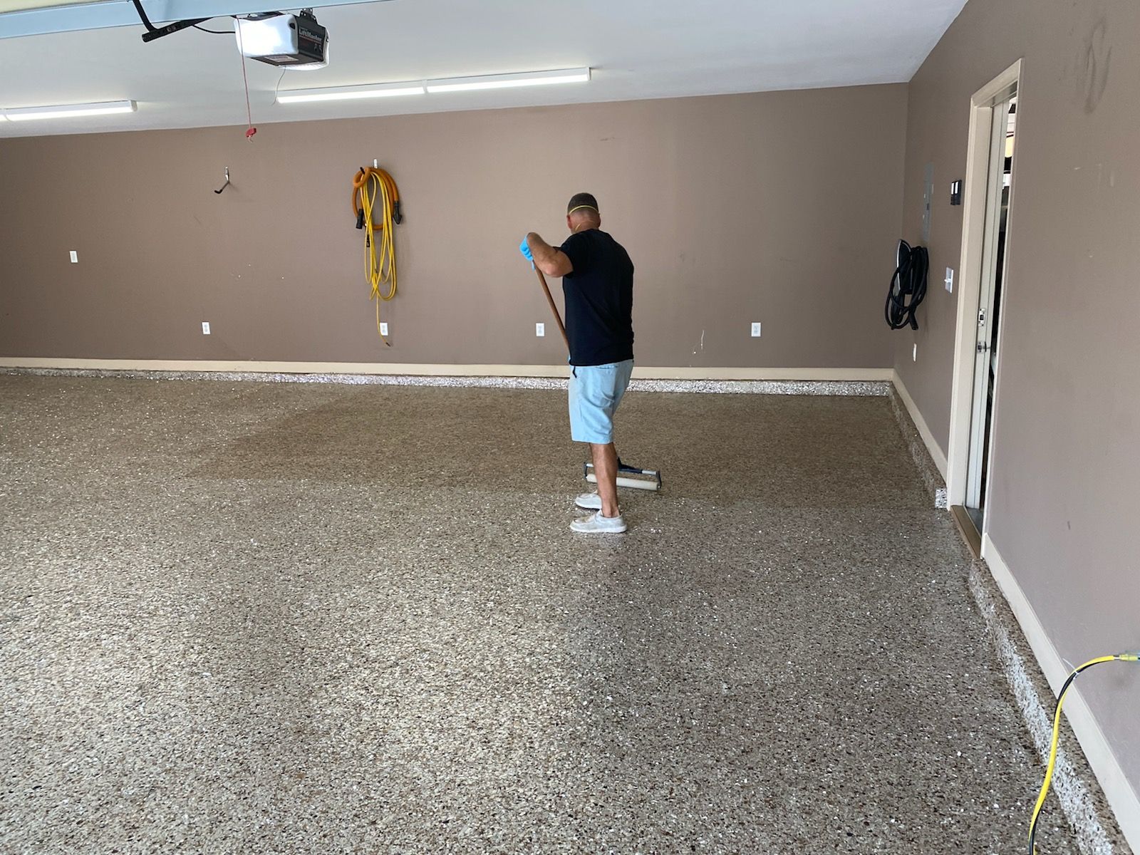 Man standing in a beige garage, holding a phone near a lift gate and scattered tools on the wall.