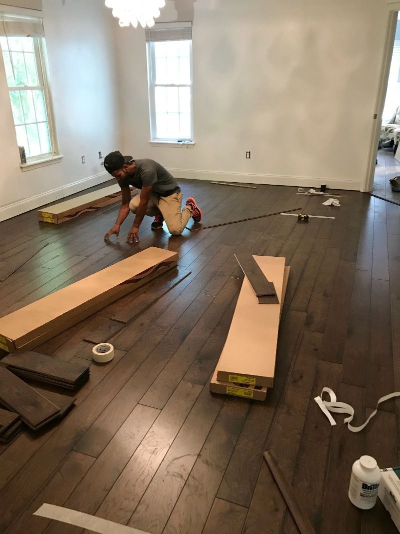 Person laying dark hardwood floors in a bright, empty room with planks, tools, and materials spread out.