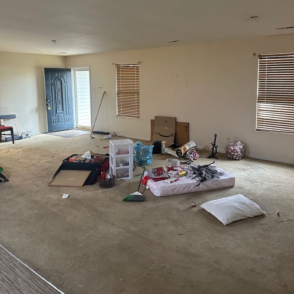 Messy empty room with scattered moving boxes, bags, and a broom on a carpeted floor