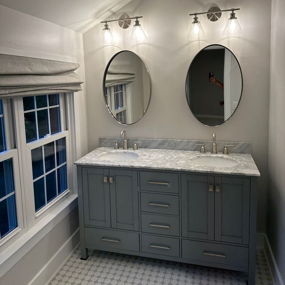 Bathroom vanity with gray cabinets, marble countertop, twin oval mirrors, and wall sconces beside a window