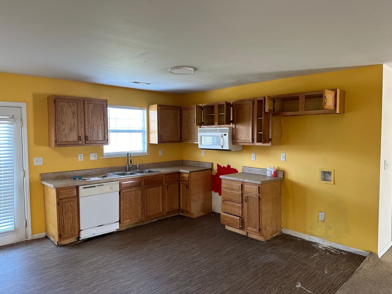 Empty yellow kitchen with wood cabinets, white appliances, and a small window and door.