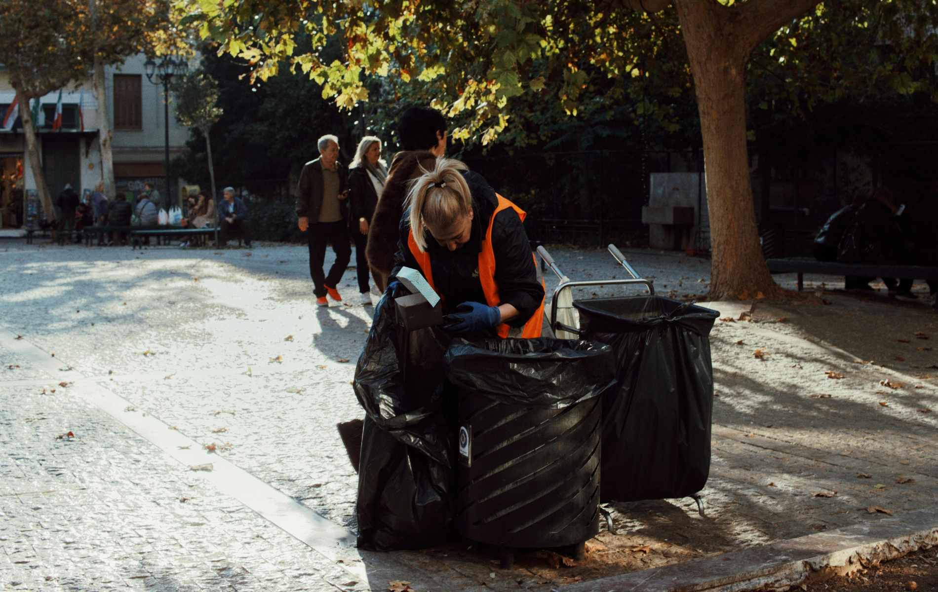 Person pushing large black garbage bags down a sunlit tree-lined street