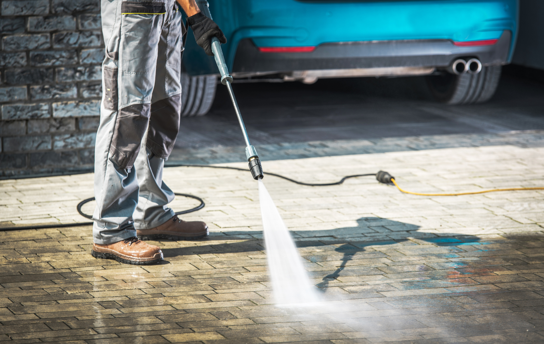 Person pressure-washing a driveway with a spray wand, water spraying near a parked blue car.