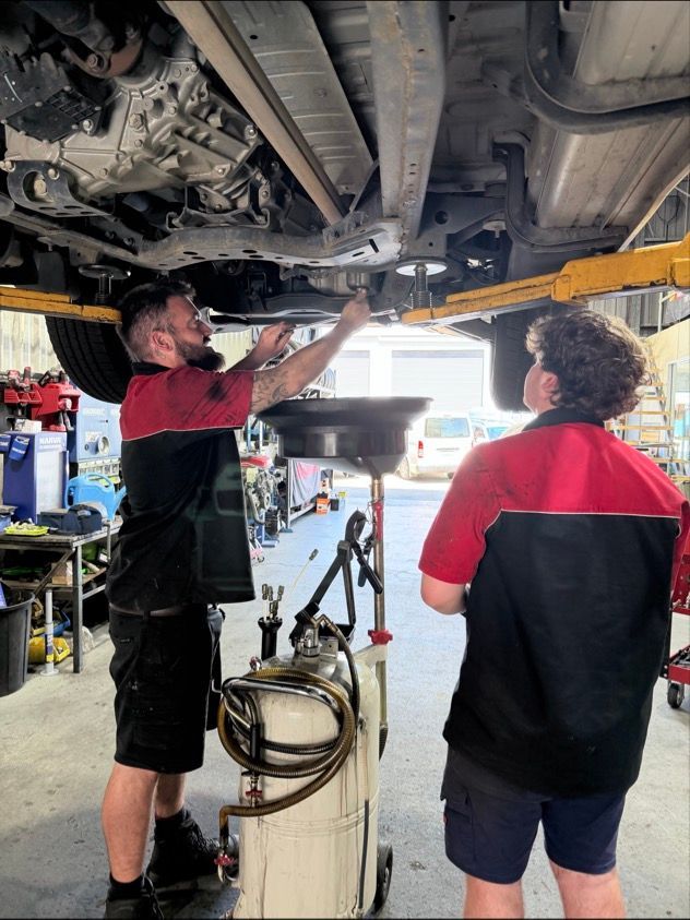 Two People in Matching Red and Black Uniforms Work Under a Raised Car — Morisset Automotive in Morisset, NSW