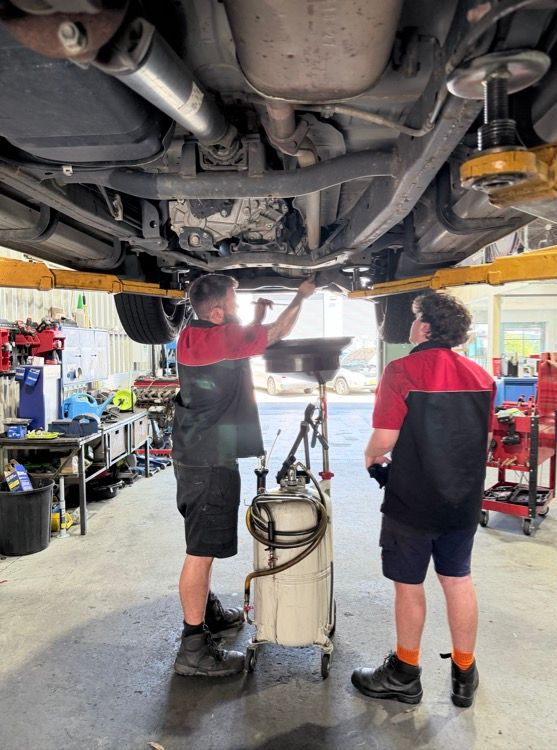 Two Mechanics in Red and Black Uniforms Work Under a Car — Morisset Automotive in Morisset, NSW