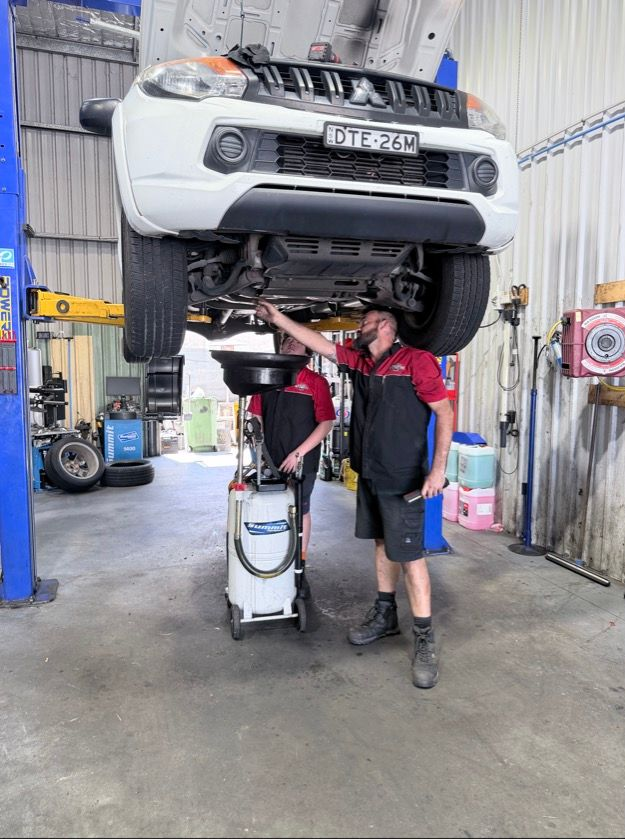 Two Mechanics in A Garage Work on The Underside of A White Vehicle — Morisset Automotive in Morisset, NSW