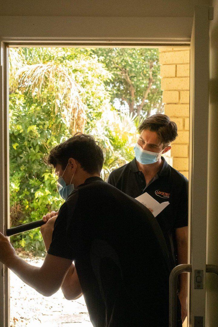 Two people wearing masks at an open doorway. One is holding a stick-like object, the other holds papers. Sunlight streams in.