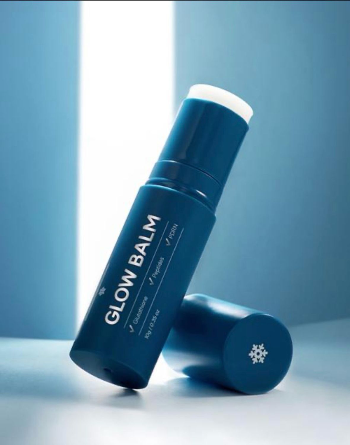 Blue Glow Balm stick with white cap on a light blue background, upright beside its cap.