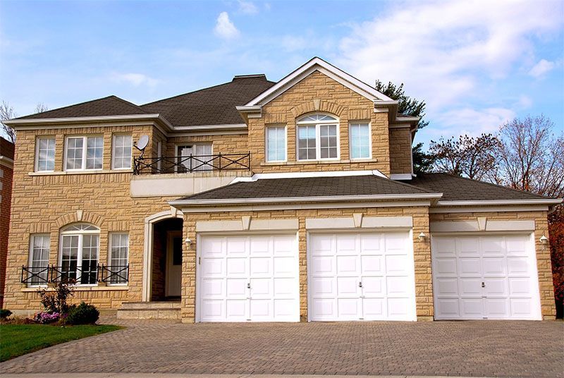 Beige brick two-story house with three white garage doors, driveway, and blue sky.