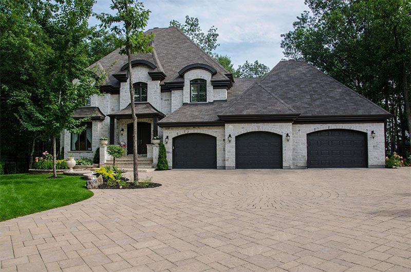 Brick house with a three-car garage and paver driveway, surrounded by trees and green lawn.