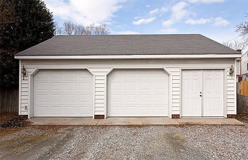 White three-car garage with two overhead doors and double doors, gray roof. Gravel driveway.