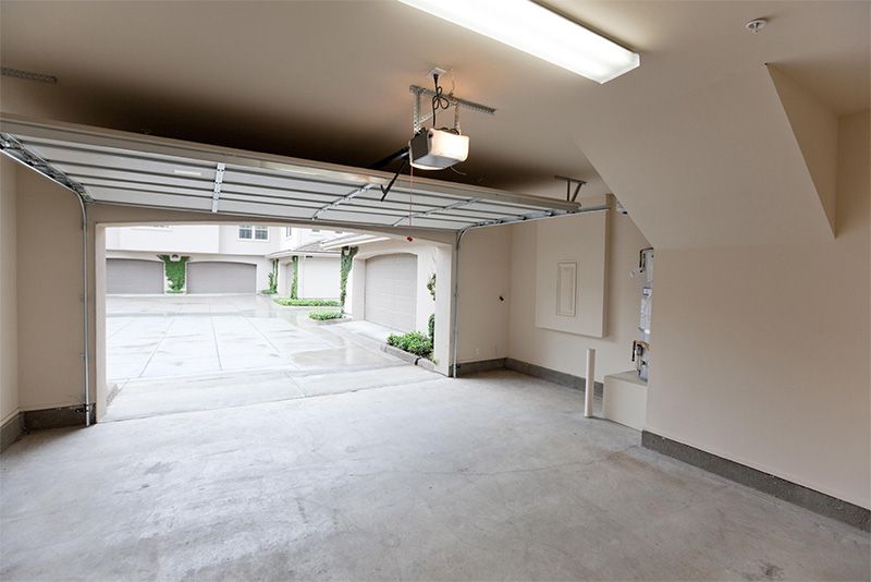 Empty garage with open door, concrete floor, white walls, and view of other garages outside.