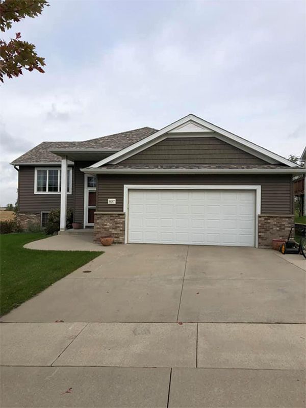 Brown-sided house with attached garage, stone accents, and a concrete driveway under an overcast sky.