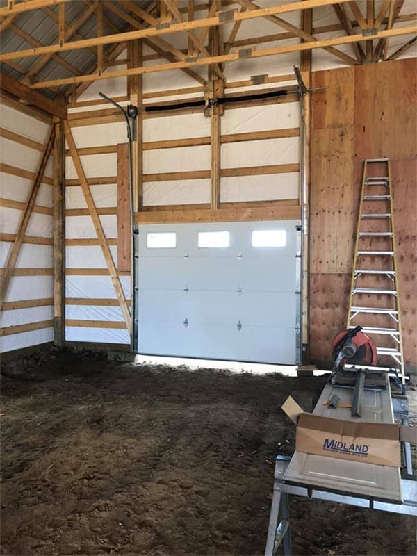 Interior view of a barn under construction with a white garage door, ladder, and wood framing.