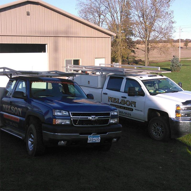 Two work trucks parked in front of a tan building. Blue truck and a white truck both have roof racks and company logos.