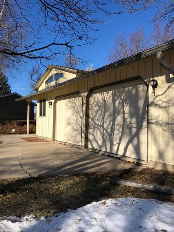 Garage with light tan exterior, two doors, and a concrete driveway. Tree shadow on the wall. Snow on the ground.