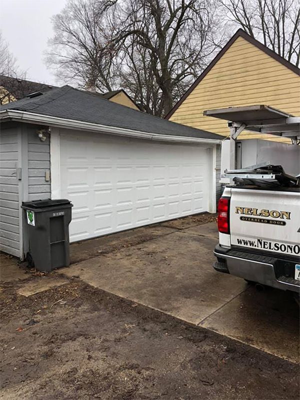 White garage with closed door next to a gray shed, with a truck parked on a concrete driveway.