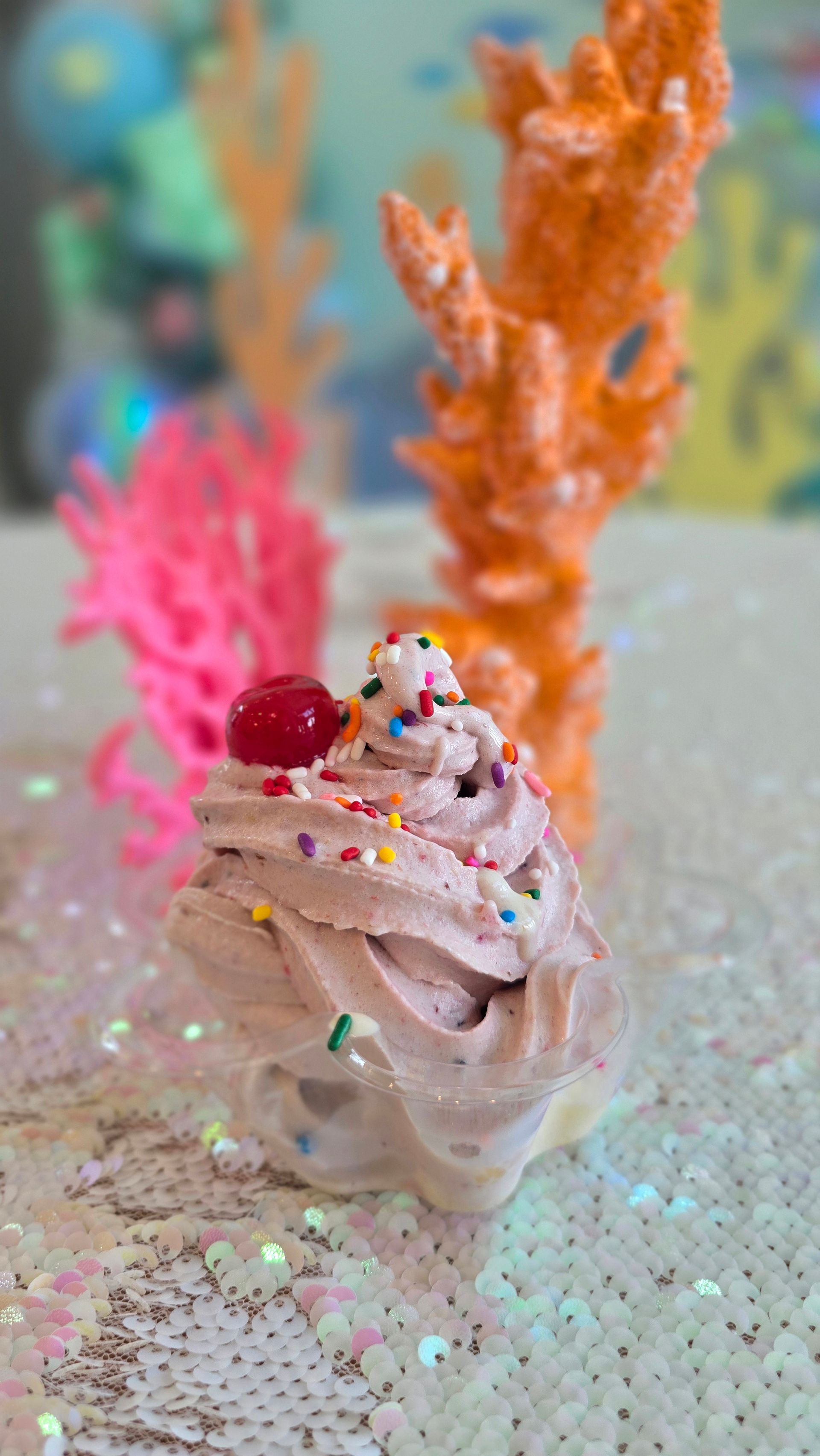 A small cup of chocolate-colored whipped cream topped with sprinkles and a cherry, set against orange and pink coral decor.