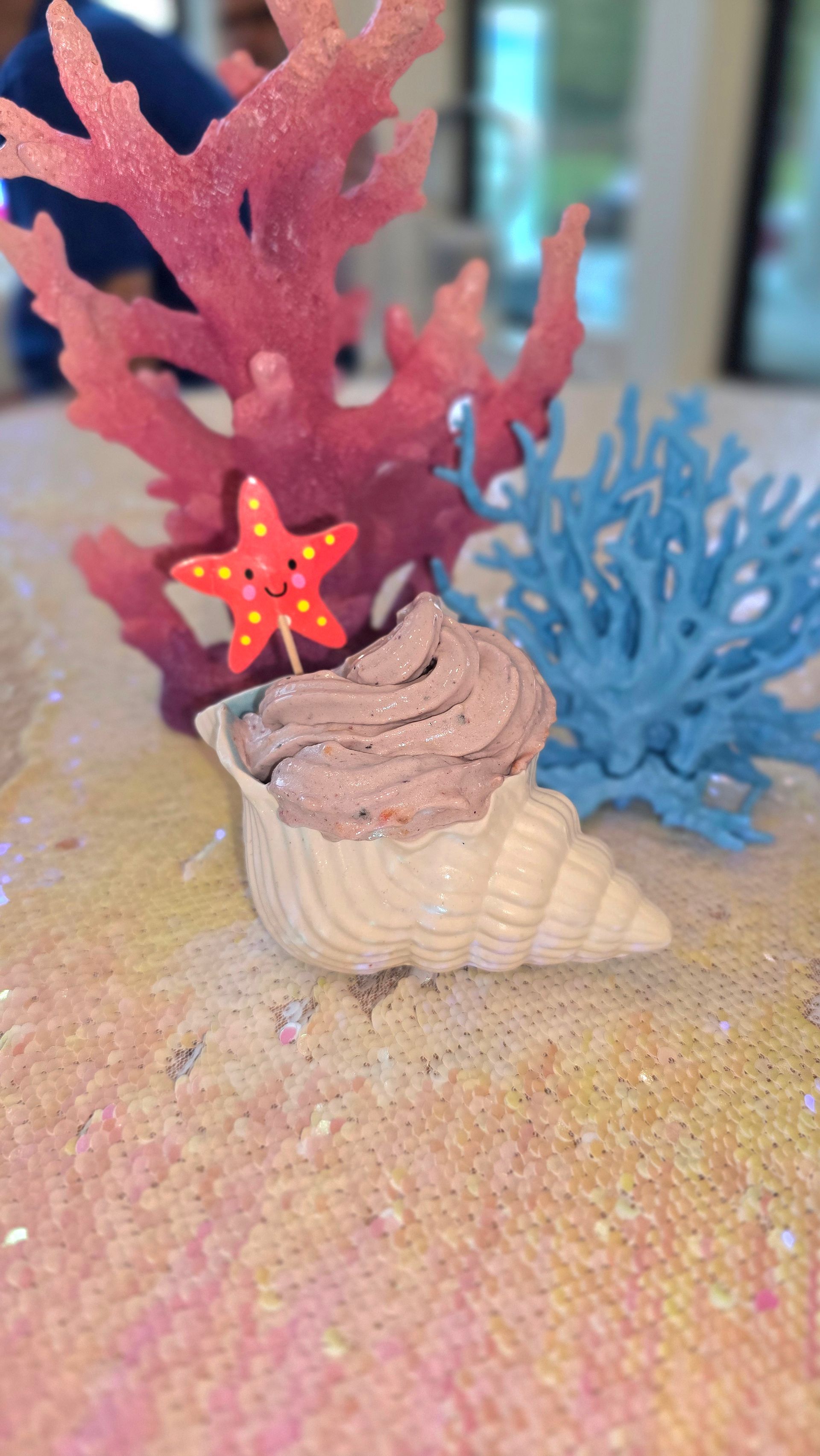 A seashell filled with chocolate mousse, topped with a smiling starfish pick, between a pink and blue coral decoration.