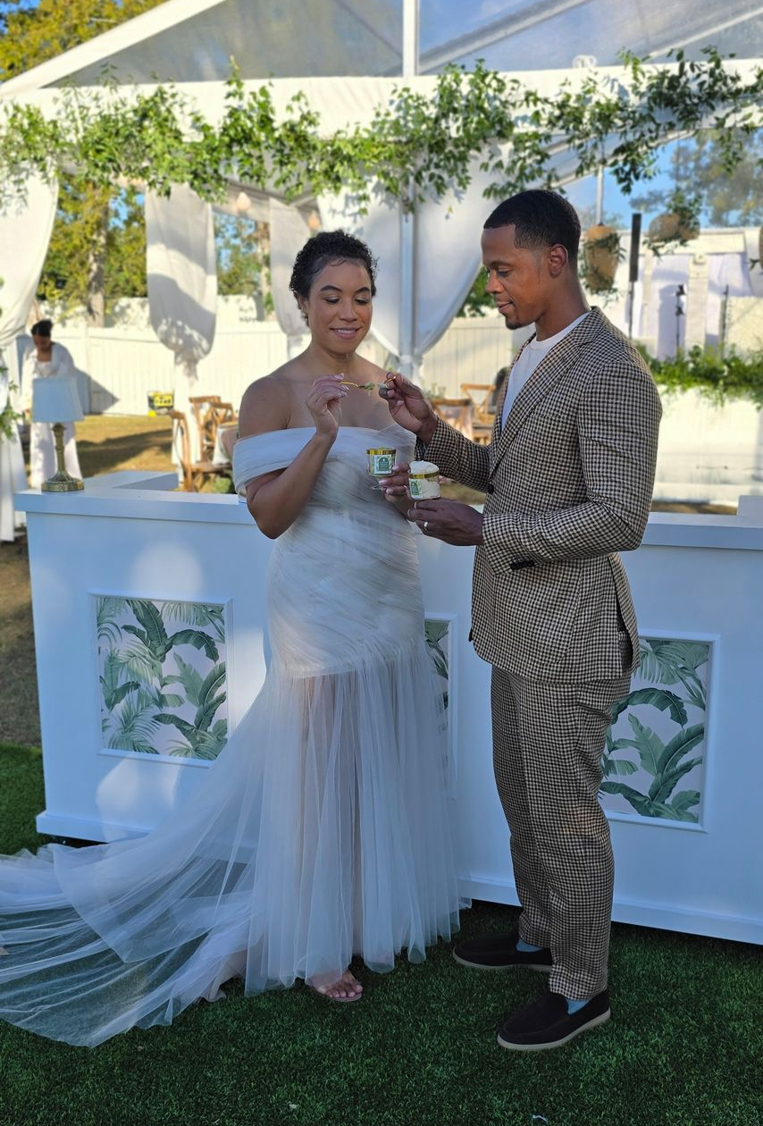 A couple stands at an outdoor bar, exchanging drinks under a tented canopy decorated with greenery and sheer fabric.