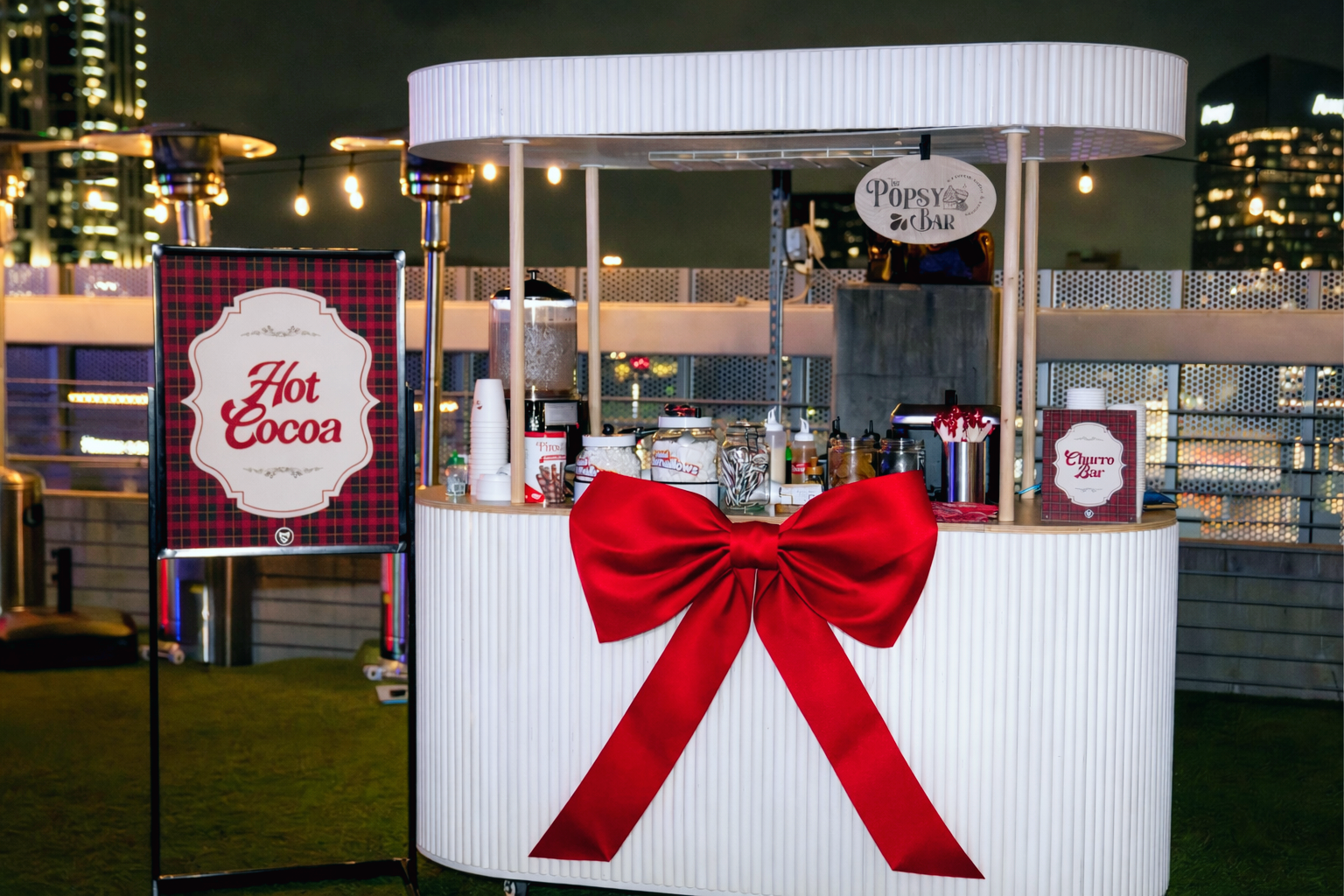 A white hot cocoa cart with a large red bow, decorated for holidays on an outdoor rooftop at night.