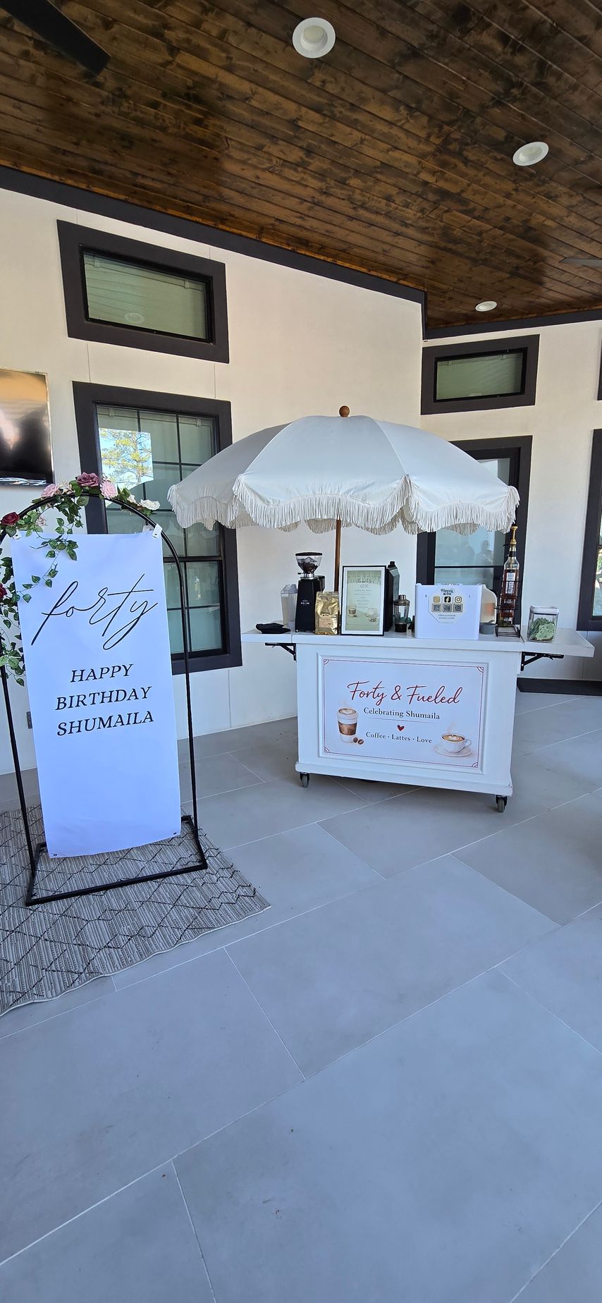 A white event cart with a matching fringed umbrella stands on a stone patio next to a welcome sign draped in greenery.