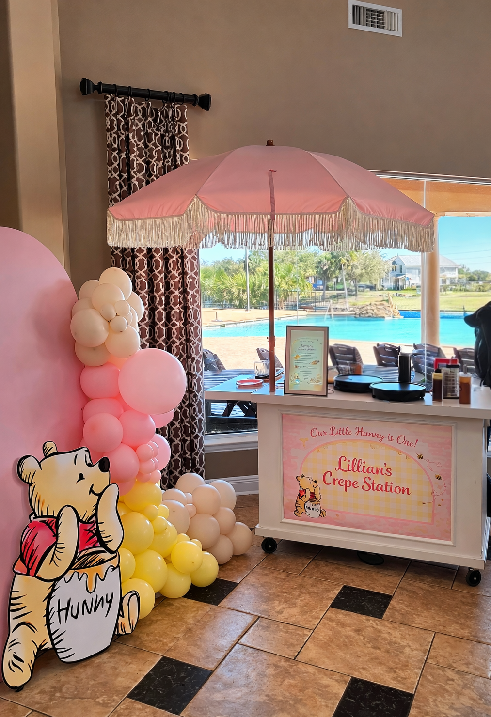 A Winnie the Pooh-themed party setup with a balloon arch, a cutout of Pooh, and a cart under a pink umbrella.