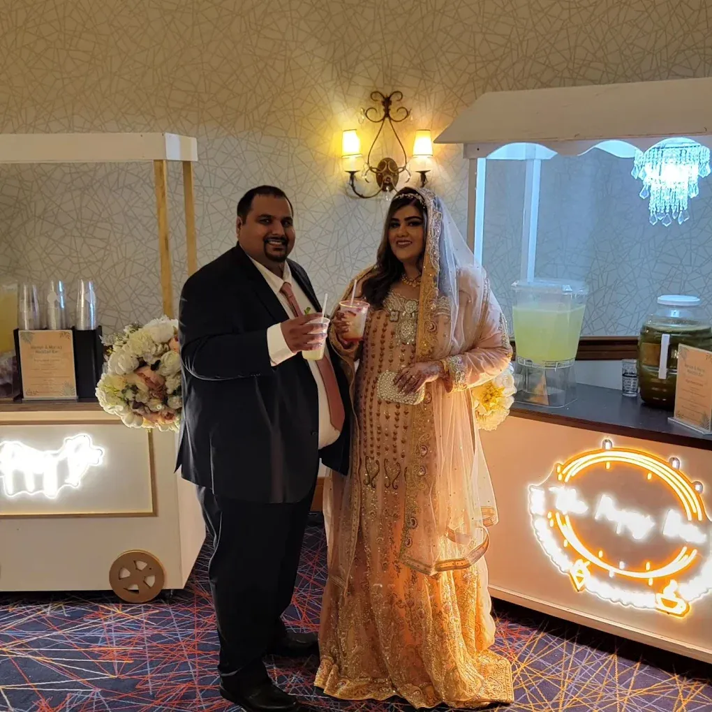 Couple poses by a cart with drinks. The woman wears a peach gown and veil. The man is in a suit, both smiling.