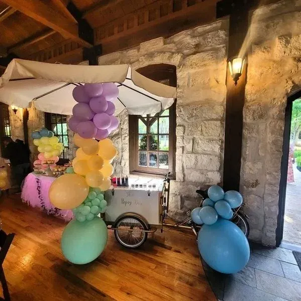 A decorative ice cream cart with pastel balloons indoors. Stone wall and wooden floor.