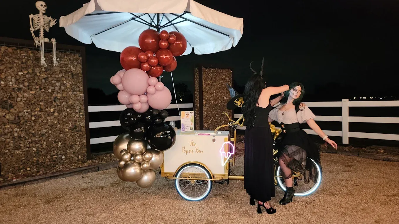 Two women pose by a decorated ice cream cart at night; balloons, a skeleton, and glowing lights create a festive atmosphere.
