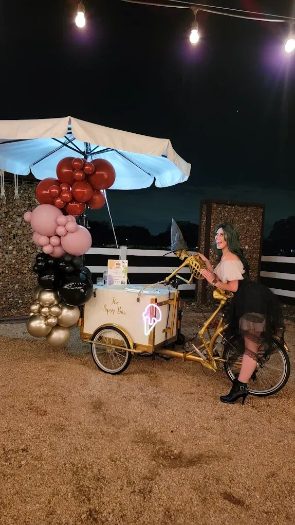 Woman riding a yellow bicycle cart decorated with balloons and carrying a cake. Outdoor setting at night.