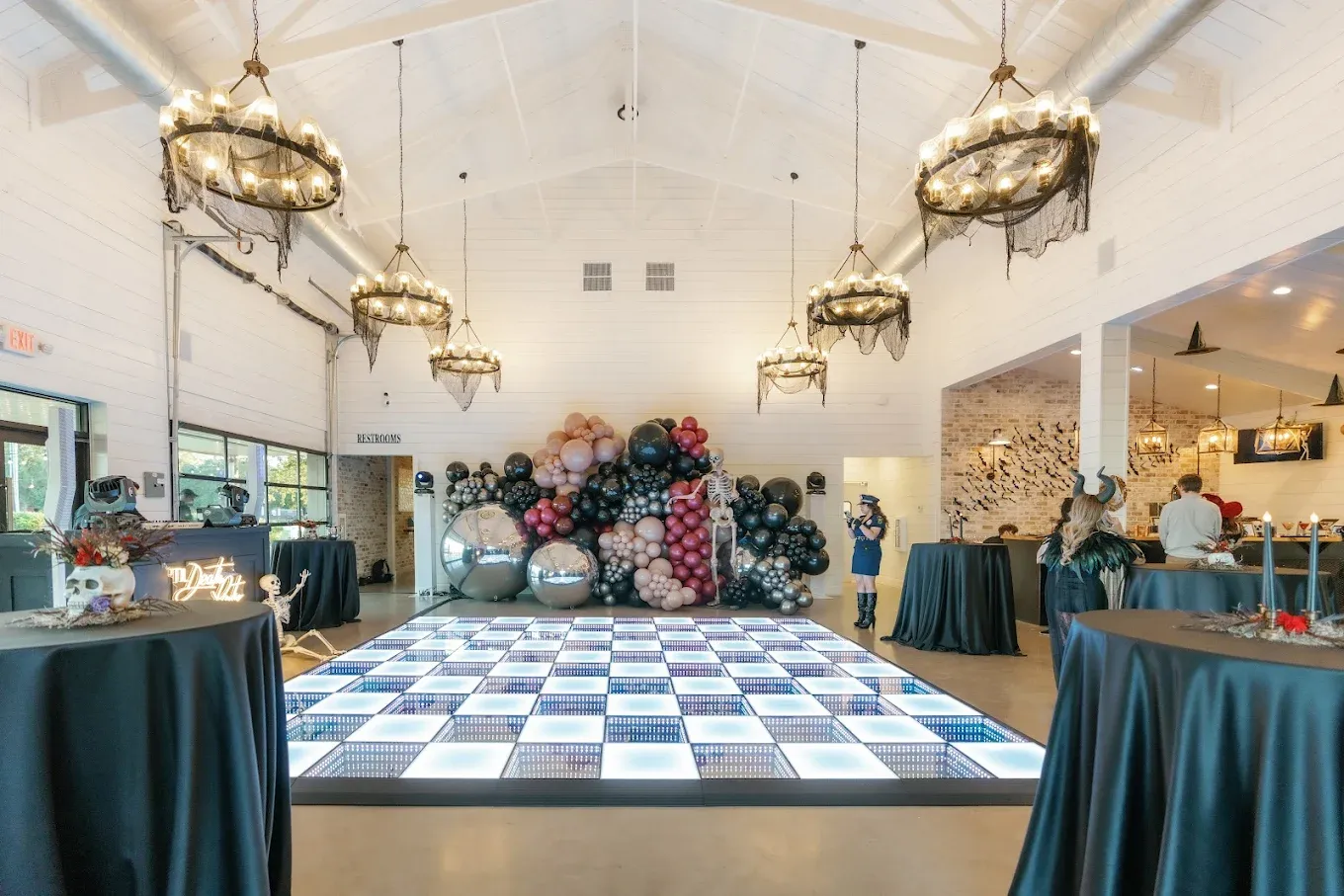 Indoor event space with a lit dance floor, tables with dark linens, and balloon backdrop.