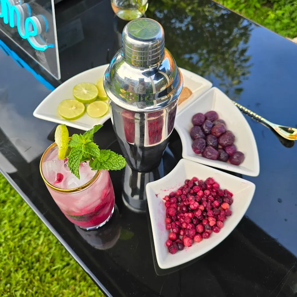 Cocktail-making setup: shaker, cocktail, lime slices, berries in white dishes on a black surface outdoors.