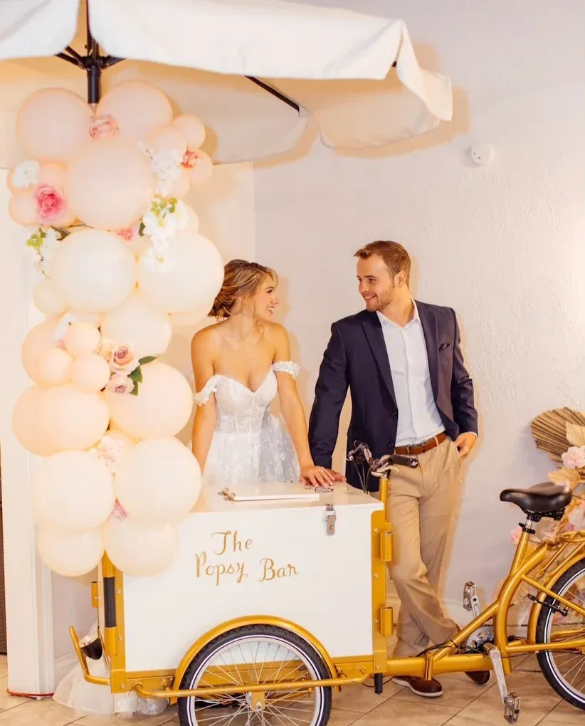 Bride and groom smile at each other, standing by a gold-trimmed cart with balloons.