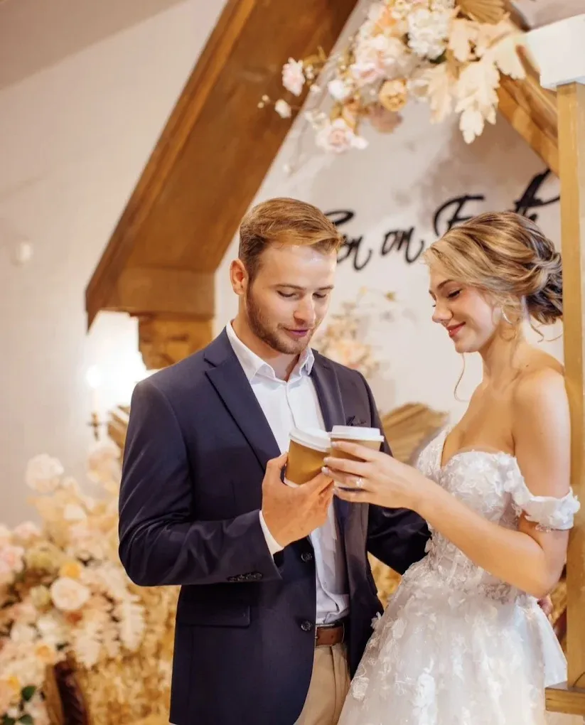 Bride and groom share coffee during their wedding. He holds the cup, she smiles in her white dress, floral decor.