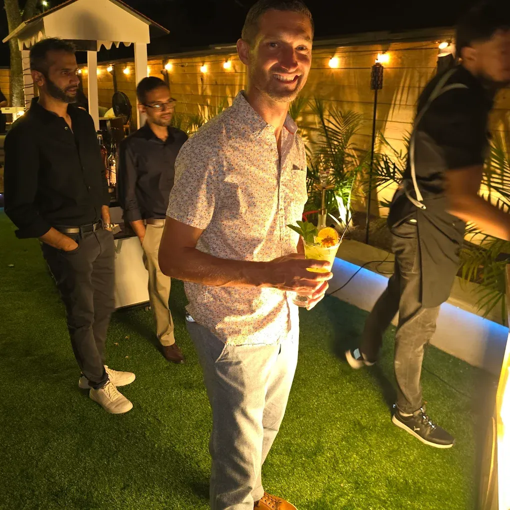 Man smiles, holding cocktail outside. Others stand by bar.